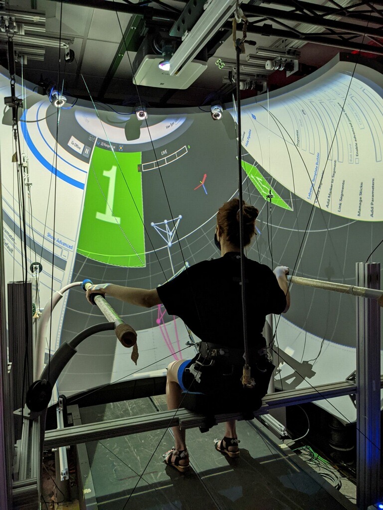 Rear view of Kristi Lyn Eaton participating in a robotics research study at Columbia University's ROAR Lab. She is secured in a body-weight support harness while holding parallel bars, facing a large projected visualization used for gait analysis, rehabilitation, and human movement research.
