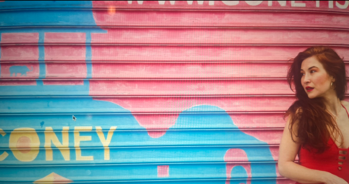 Portrait of Kristi Lyn Eaton standing in front of a painted metal storefront mural with bright blue and pink abstract shapes and partial lettering. She is wearing a red top, has long auburn hair over one shoulder, and is looking to the right.
